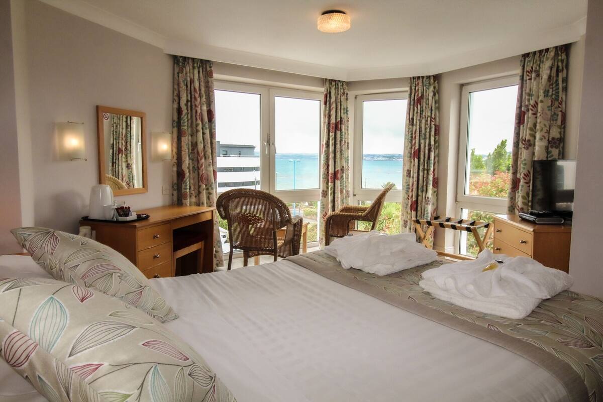 A hotel room with a bed, two chairs, a desk, a television, and large windows with curtains.