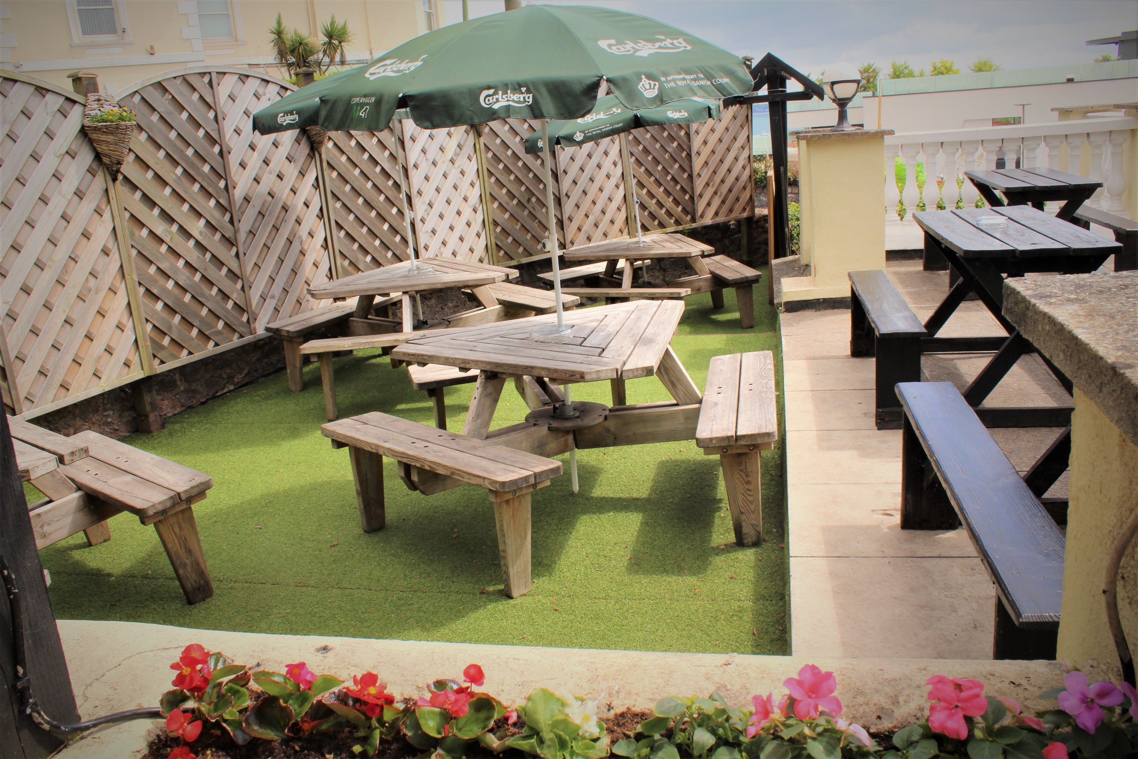 A patio area with wooden tables and benches, green artificial grass, and a lattice fence.