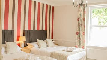 Hotellihuone, jossa on kaksi sänkyä, raidallinen tapetti, kattokruunu ja ikkuna, jossa on verhot.