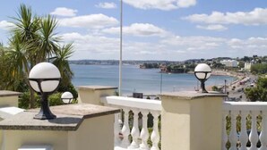 A balcony with a white balustrade, a flagpole with a flag, and a view of a coastal town. - The Heritage Hotel (Torquay)