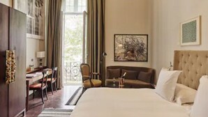 A hotel room with a large bed, a desk, a chair, and a sofa. There is a window with curtains, a painting on the wall, and a door leading outside. - Hotel B (Lima)