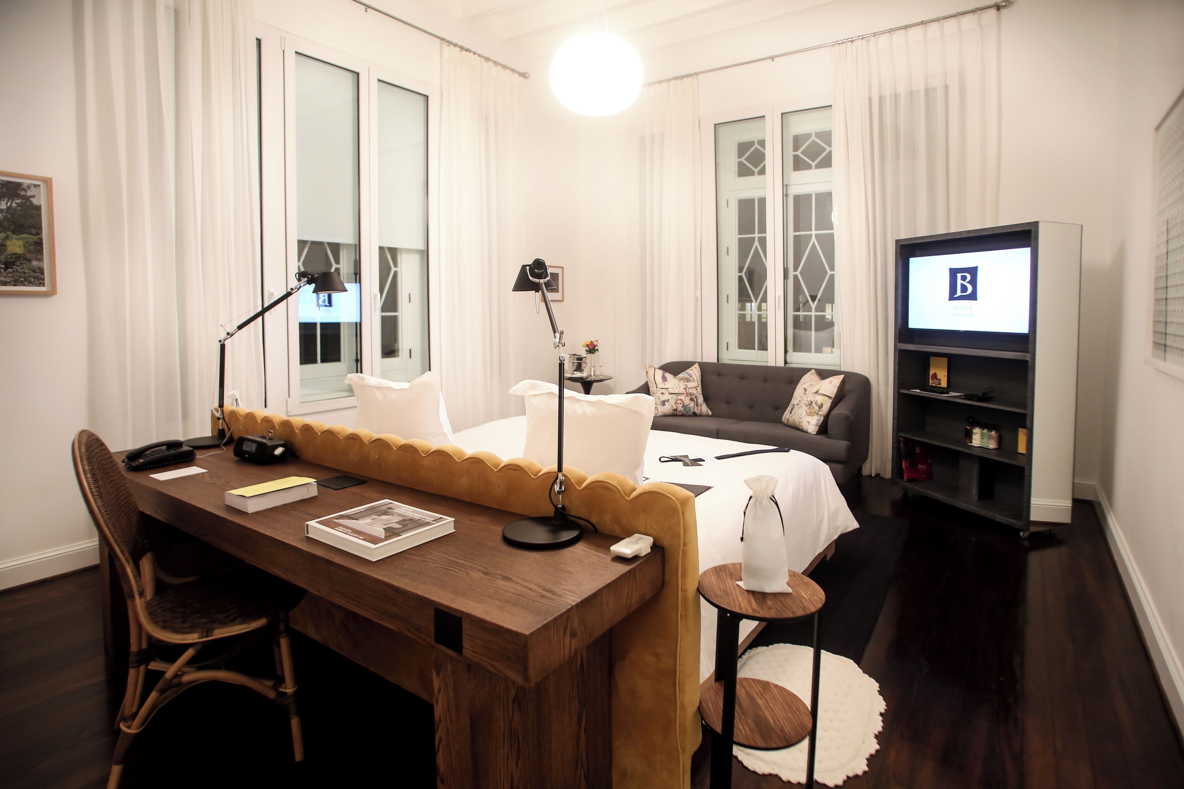 Une chambre d’hôtel moderne avec un bureau en bois, un canapé confortable, un lit avec une literie blanche, une télévision et une bibliothèque.