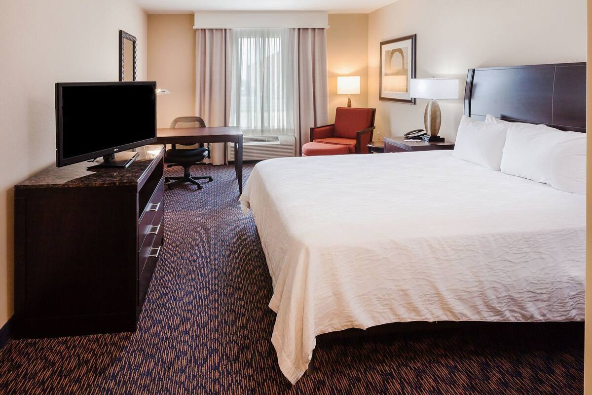 A hotel room with a bed, a desk with a TV, a chair, a lamp, and a window with curtains.