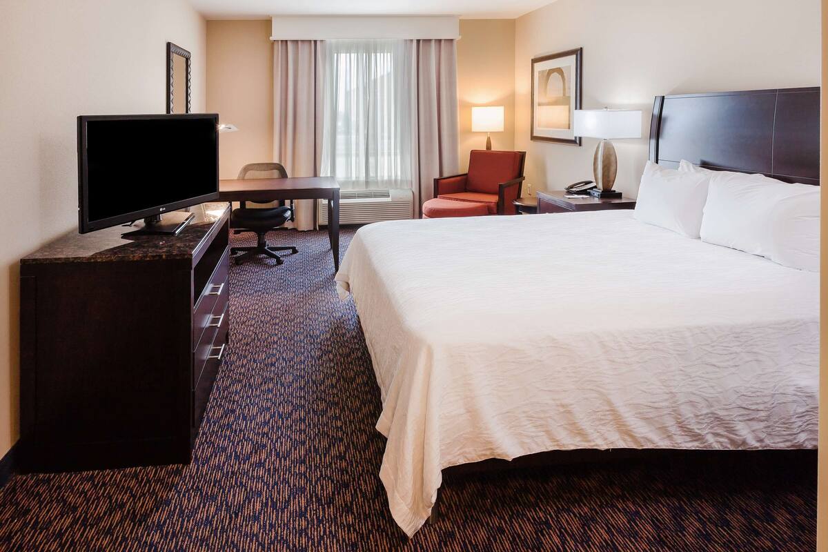 A hotel room with a bed, a desk with a computer, a chair, a TV, and a lamp.
