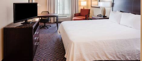 A hotel room with a bed, a desk with a computer, a chair, a TV, and a lamp.