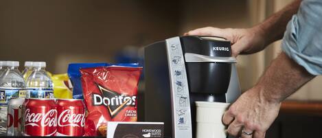 A coffee machine with a manual coffee pot, a bag of Doritos, and several cans of Coca-Cola on a counter.