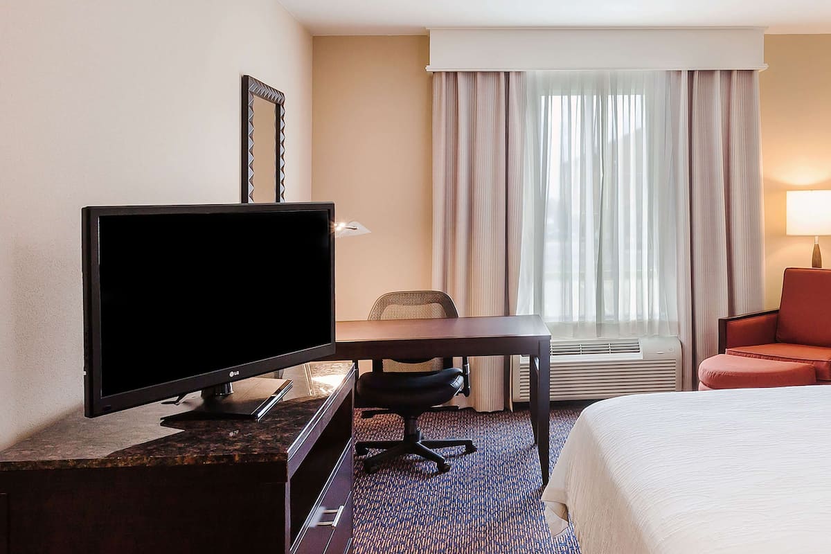 A hotel room with a bed, a desk with a television, a chair, a lamp, and a window with curtains.