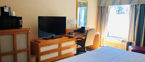 A hotel room with a bed, a desk with a television, a chair, a lamp, and a window with curtains.