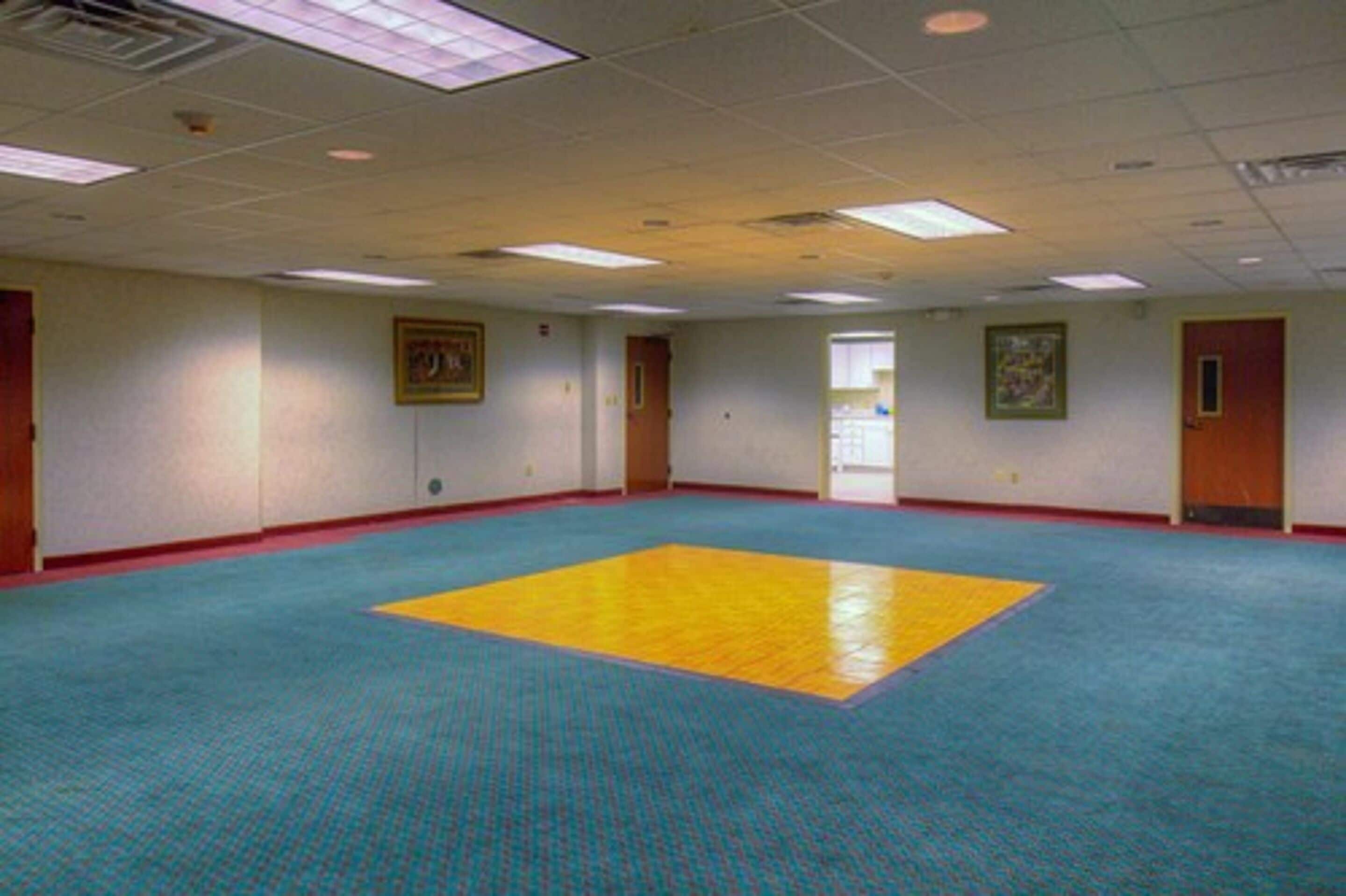 A large, empty room with a blue carpet, a yellow and blue floor mat, and white walls with framed pictures.