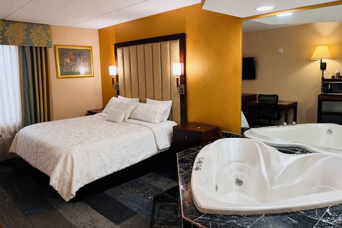 A hotel room with a bed, a sitting area with a chair, a desk, a large bathtub, and a television.