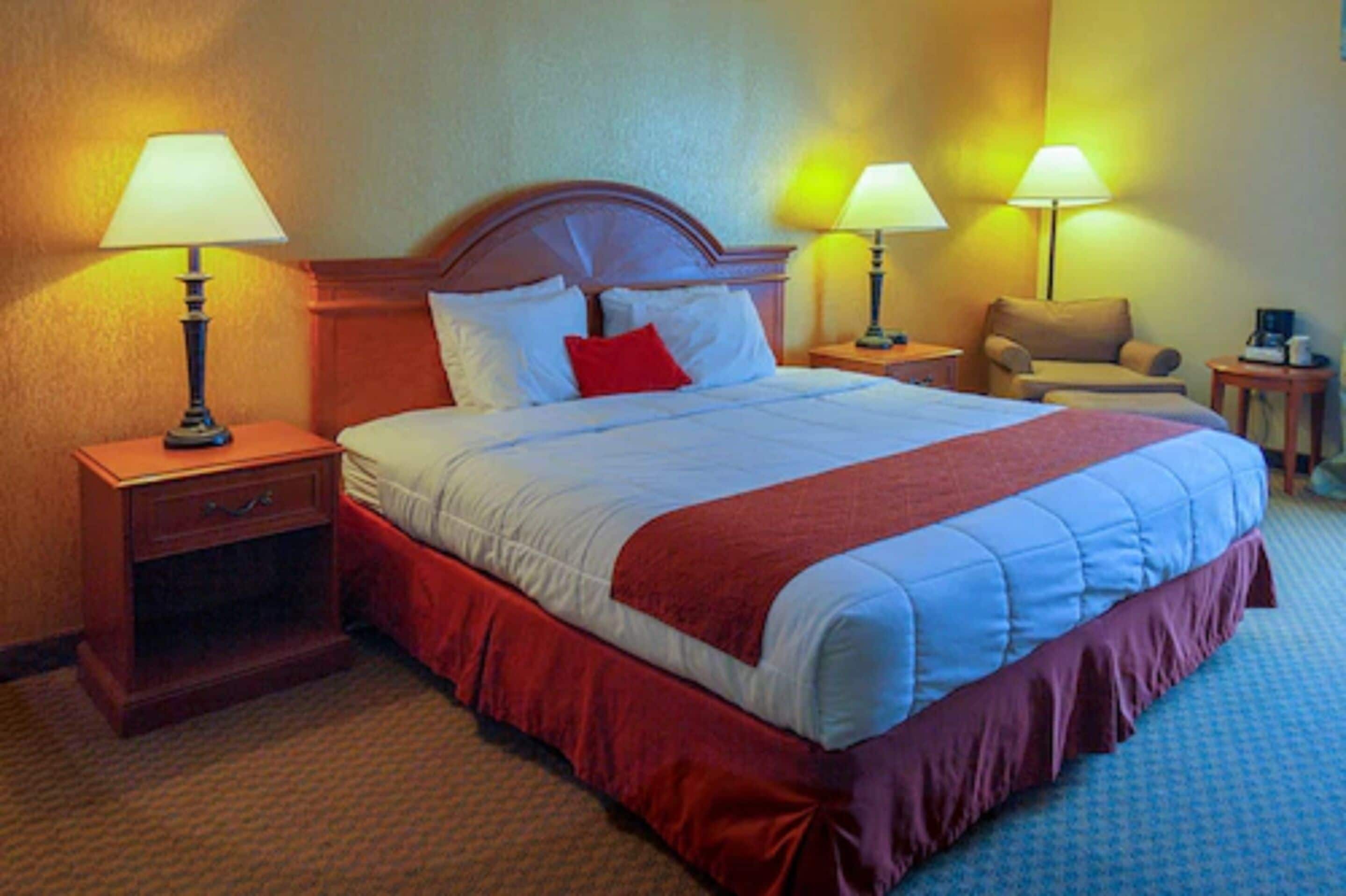 Een hotelkamer met een groot bed, twee bedlampen, een nachtkastje met lamp, een stoel en een kleine tafel.