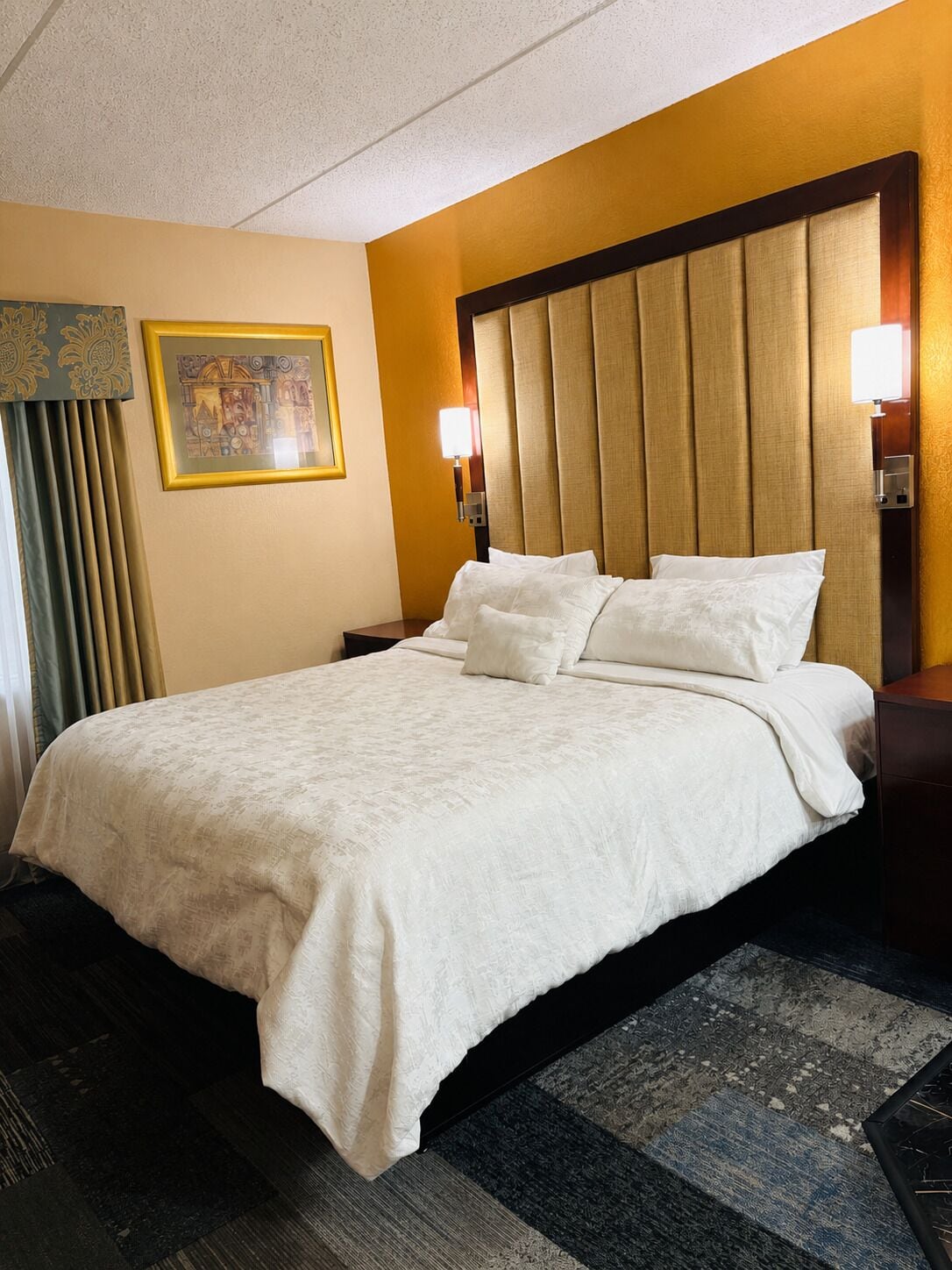A hotel room with a large bed, a headboard, two bedside lamps, a framed picture on the wall, and a window with curtains.