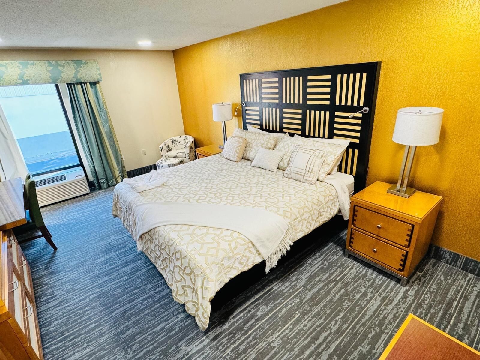 A hotel room with a large bed, a bedside table with a lamp, a chair, and a view of the sea through a window.