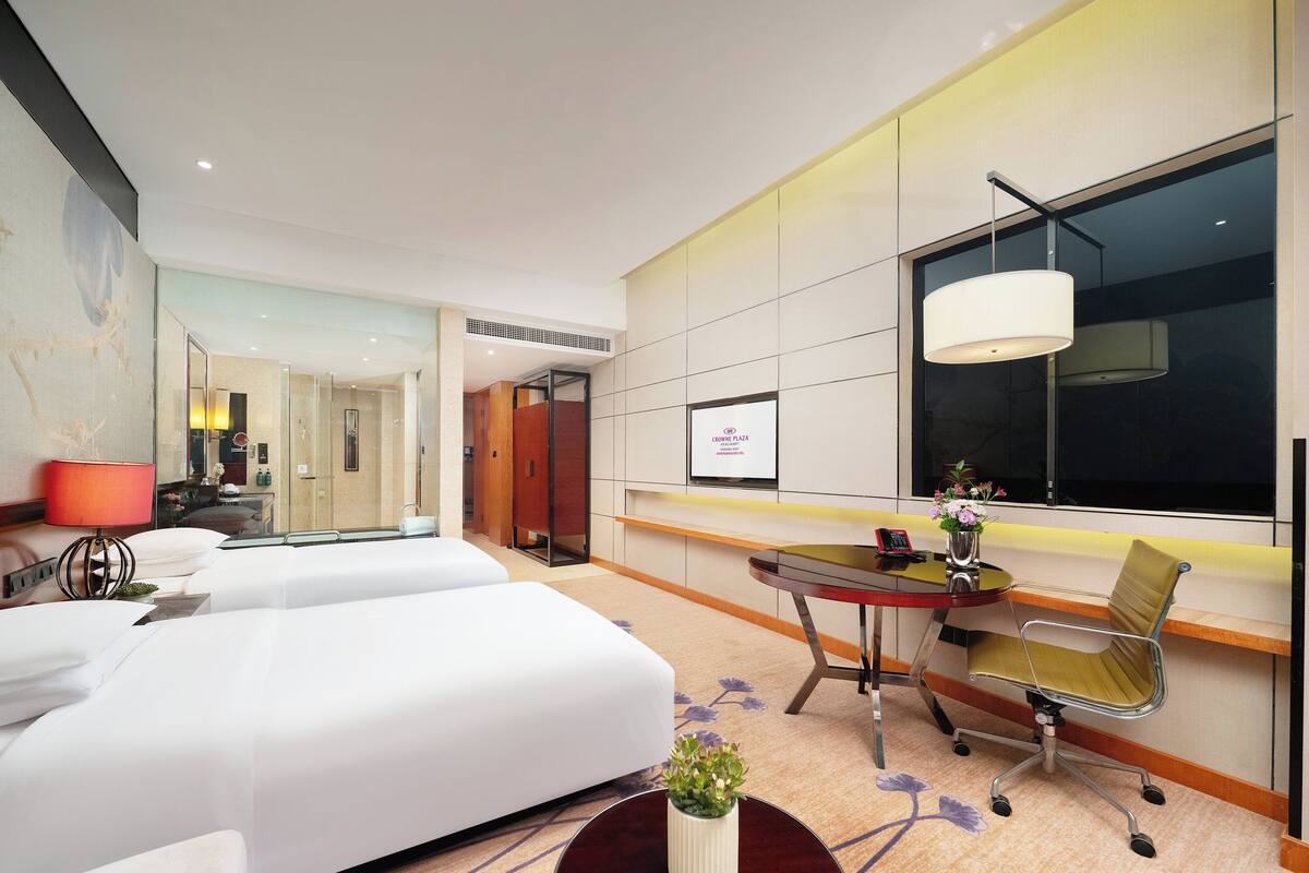 A modern hotel room with two beds, a desk, a chair, a lamp, and a large window.