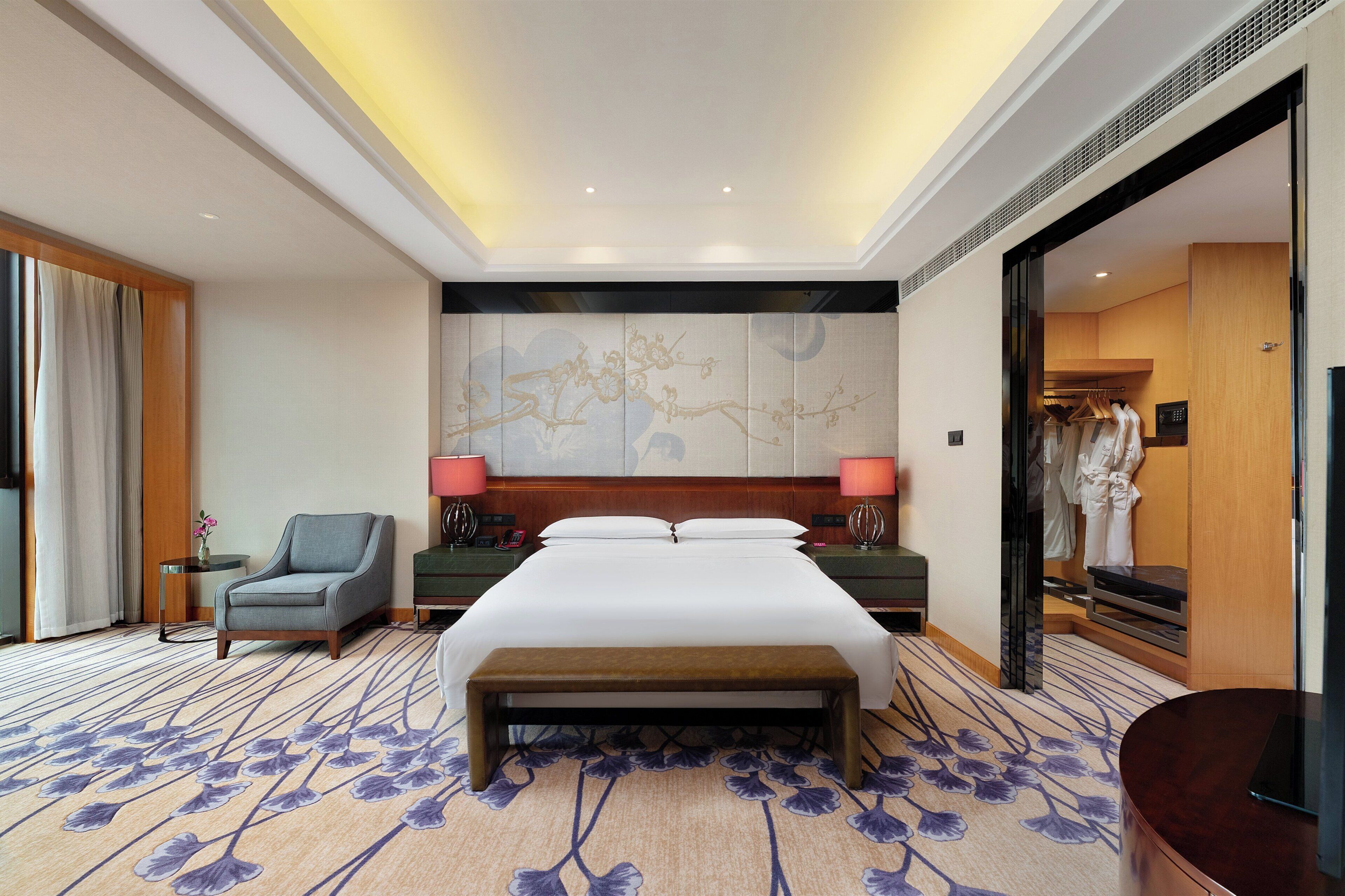 A hotel room with a large bed, a bedside table with lamps, a chair, a bench, and a large mirror.