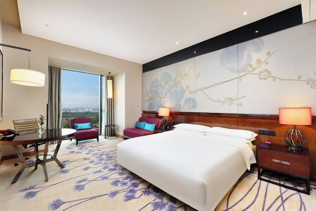 A hotel room with a large bed, a desk with a chair, a sofa, and a view of the city.