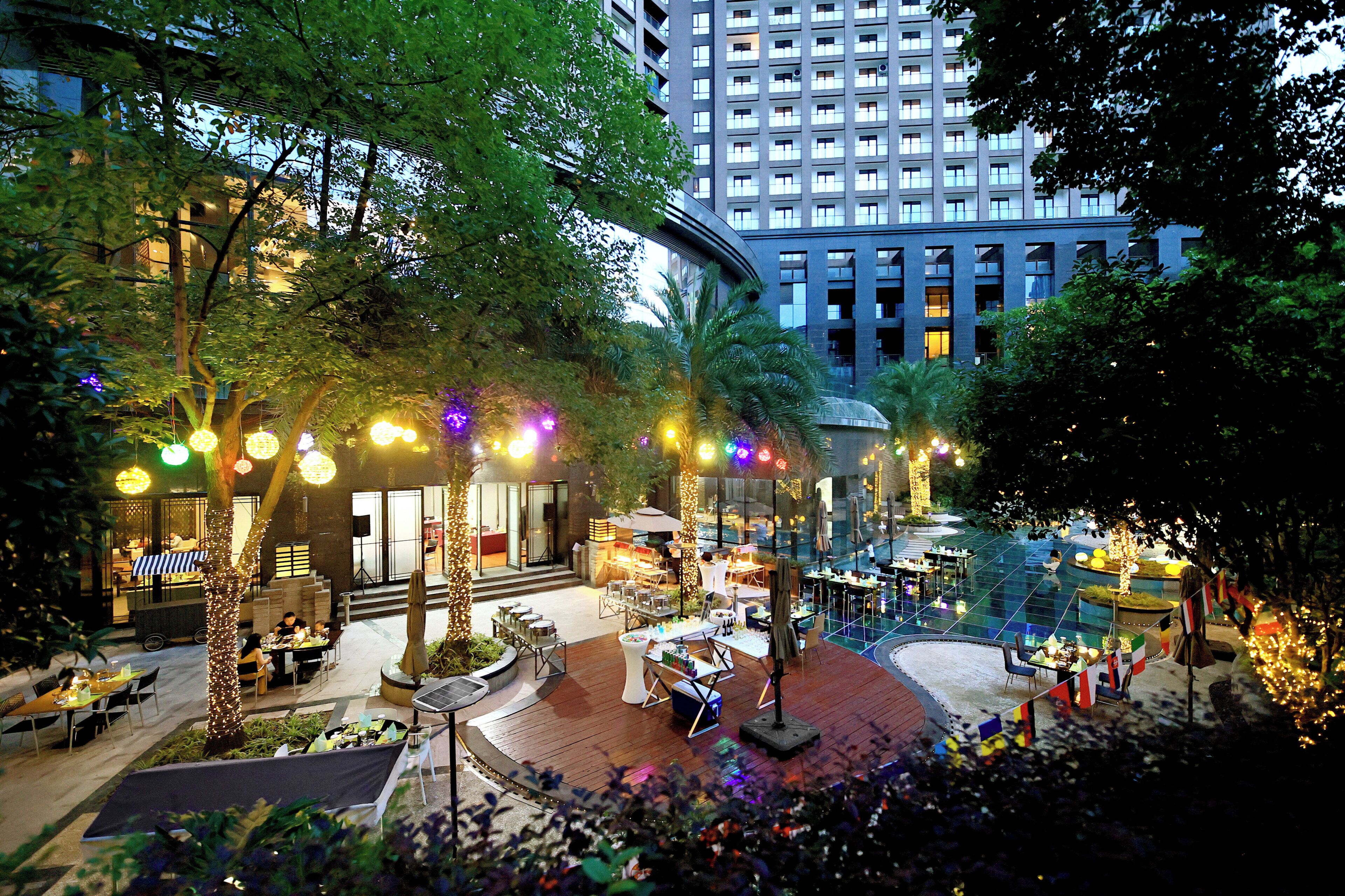 An outdoor dining area with tables and chairs, surrounded by trees and illuminated by string lights.