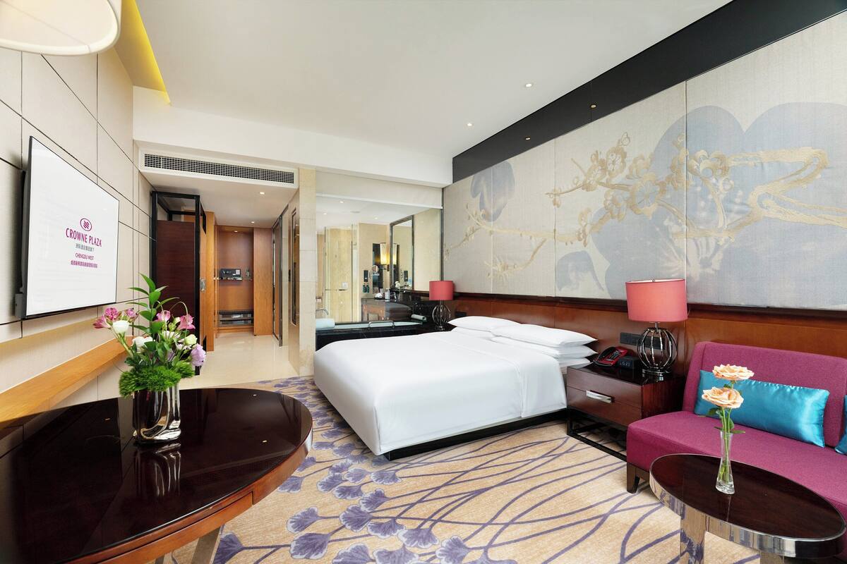 A hotel room with a bed, a desk, a sofa, a TV, and a large artwork on the wall.