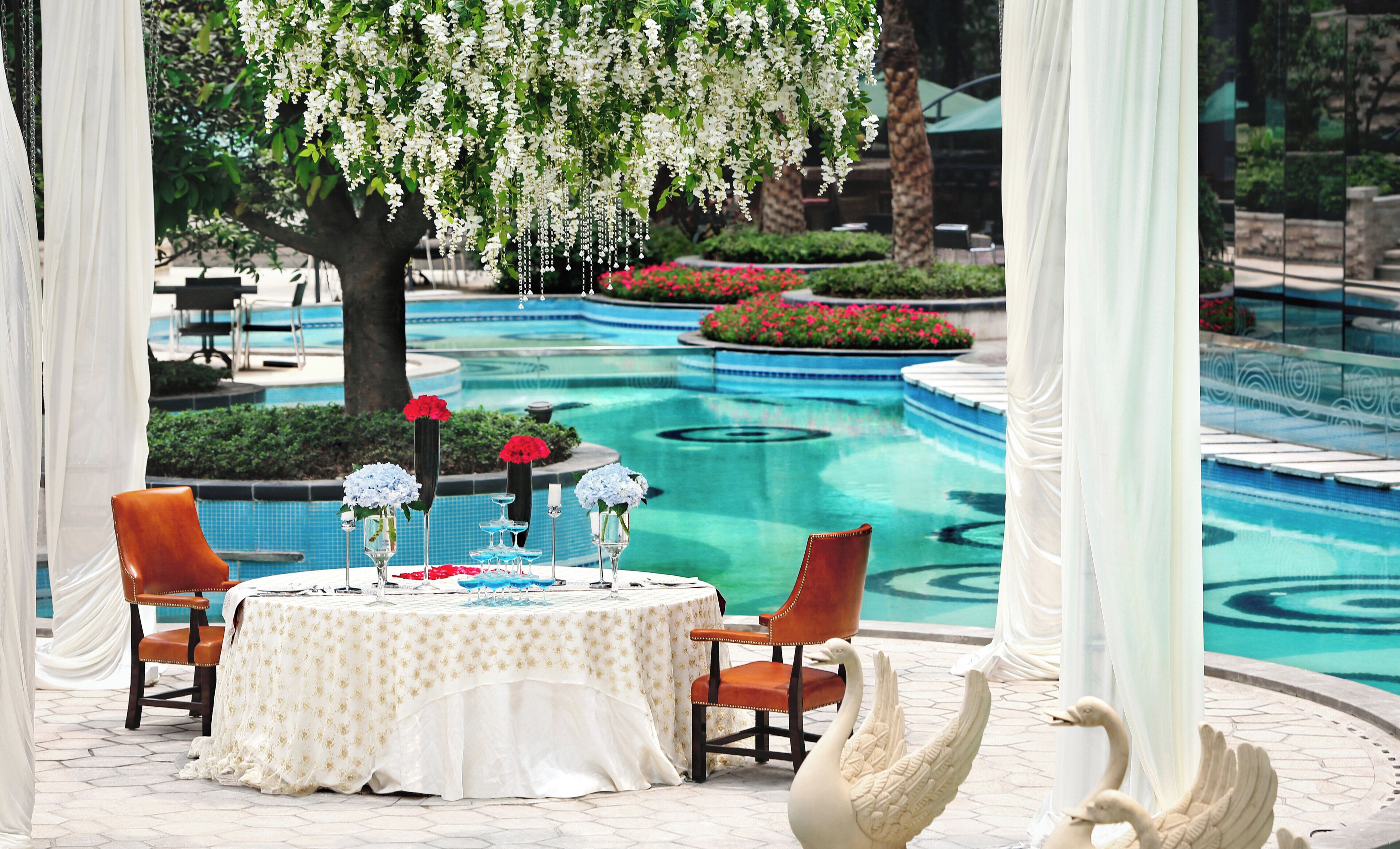 A dining area with a pool view, featuring a table set with white tablecloth, chairs, and decorative items.