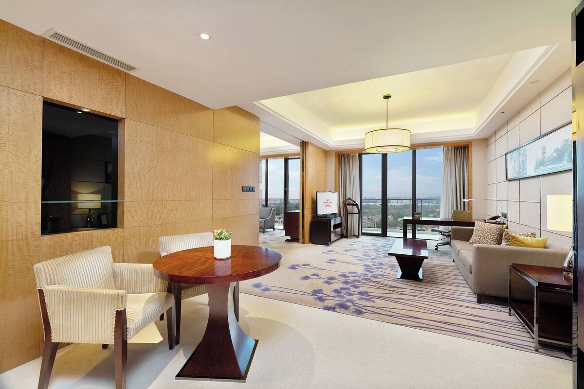 A modern hotel room with a large window, a round wooden table, a sofa, and a chair.