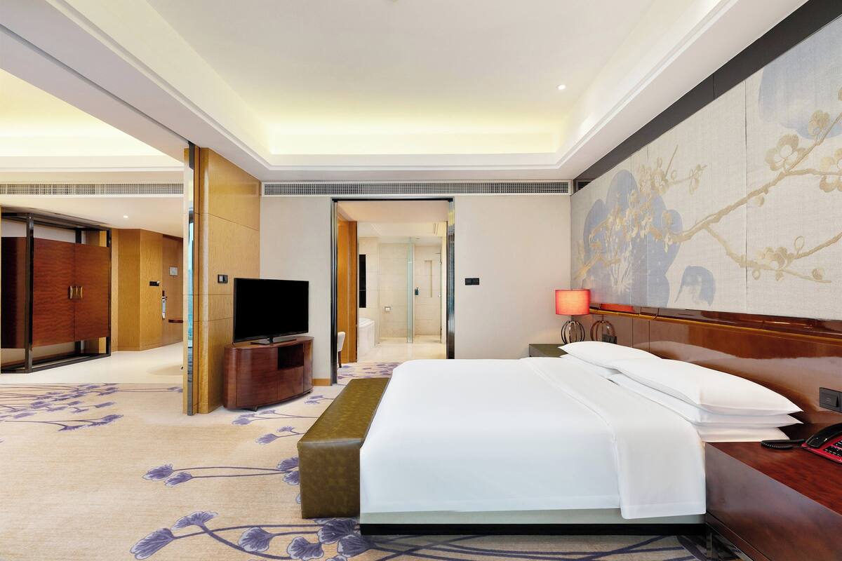 A hotel room with a bed, a television, a desk, a lamp, and a painting on the wall.