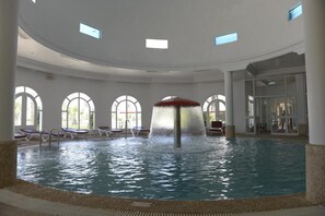 An indoor swimming pool with a central water feature, sunlit by large windows, and surrounded by lounge chairs.