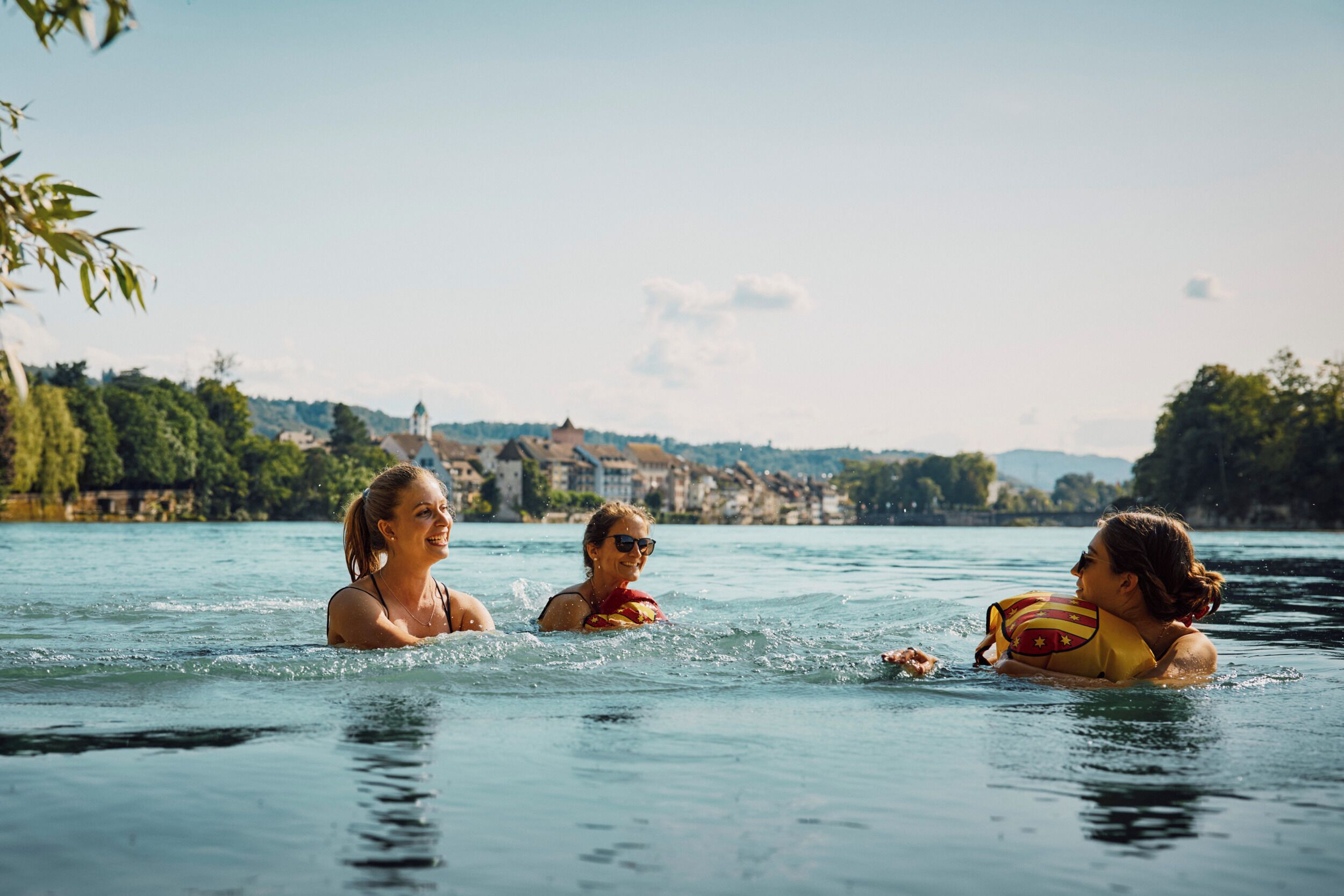 Three people in a swimming pool with a scenic view of buildings and trees in the background.