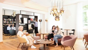 A modern lounge area with round tables, comfortable seating, and a bookshelf in the background. - Hotel Schützen (Rheinfelden)