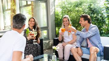 Un grupo de personas sentadas en muebles de mimbre al aire libre, disfrutando de bebidas y conversación en un espacio luminoso e inundado de sol, con grandes ventanales y follaje al fondo.