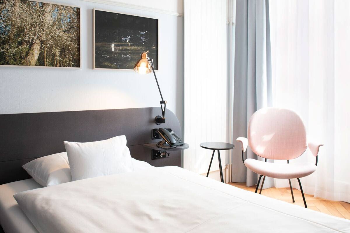 A modern hotel room with a large bed, a bedside table with a lamp, a small round table, and a pink chair. There are two framed pictures on the wall above the bed.