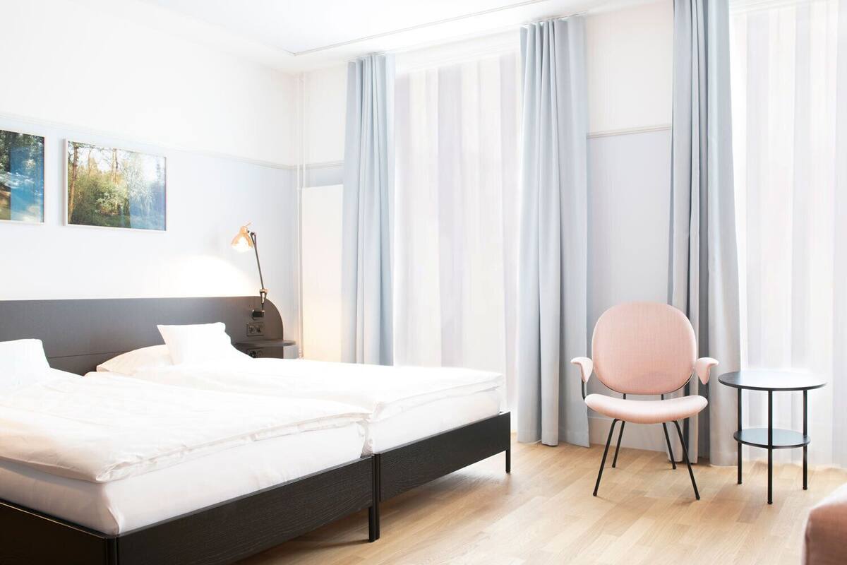 A modern hotel room with a bed, a chair, a small table, and large windows with curtains.