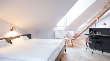 Un dormitorio moderno con una cama grande, un escritorio y una silla. El techo es inclinado y hay una ventana que proporciona luz natural.