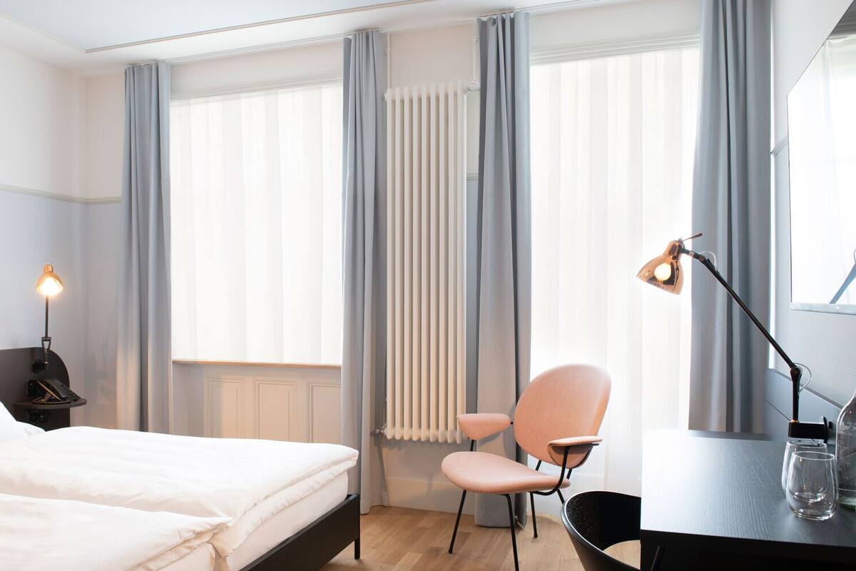A hotel room with a bed, a desk with a lamp, a chair, and a radiator.