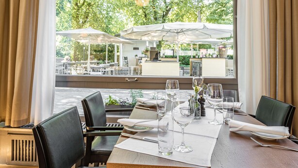 A dining table set with glasses and cutlery, a window with curtains, and outdoor seating visible through the window. - Hotel Schützen (Rheinfelden)