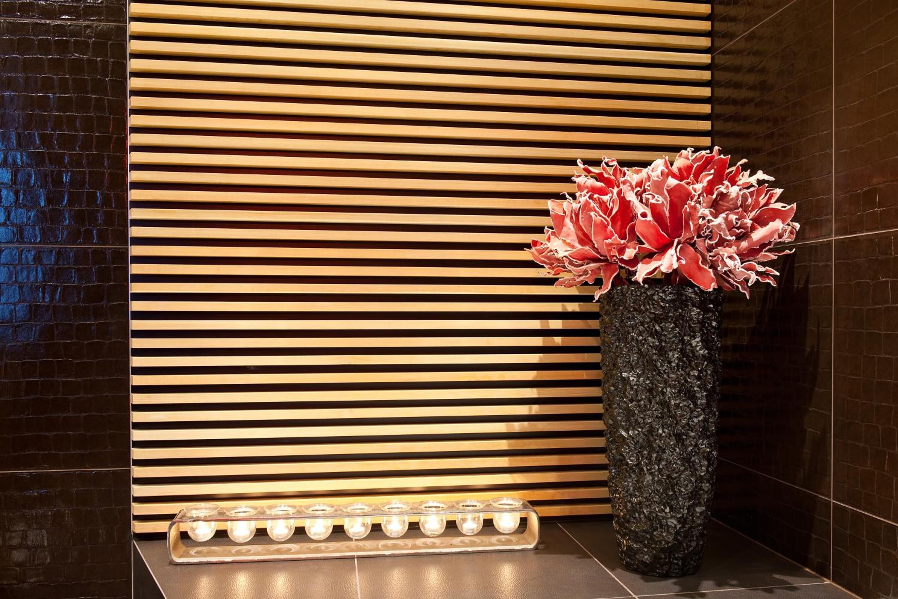 Un interior moderno con un revestimiento de listones de madera, un jarrón negro texturizado con flores rojas y una fila de luces iluminadas.