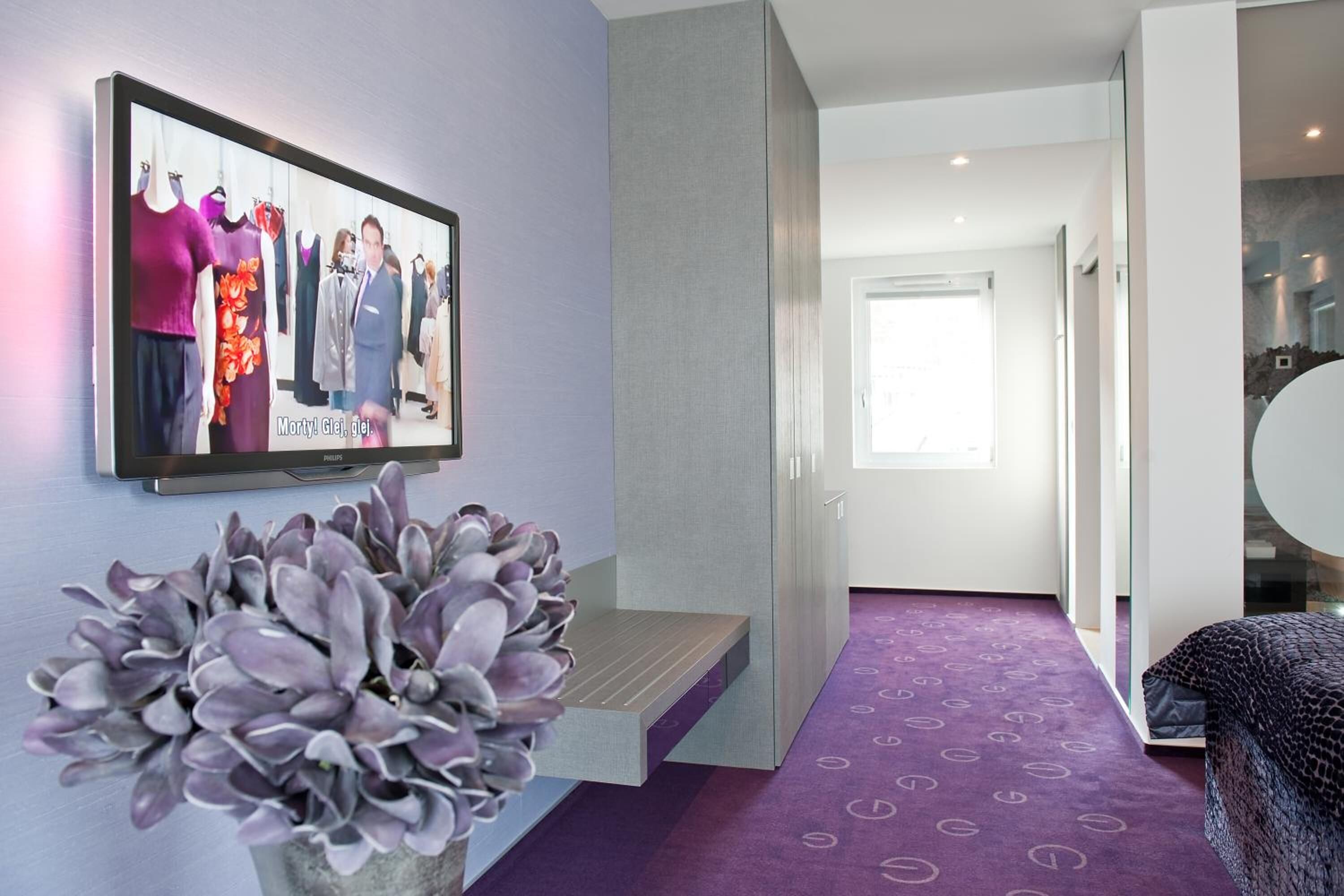 A modern hotel room with a purple carpet, a large mirror, and a wall-mounted television displaying a fashion show.