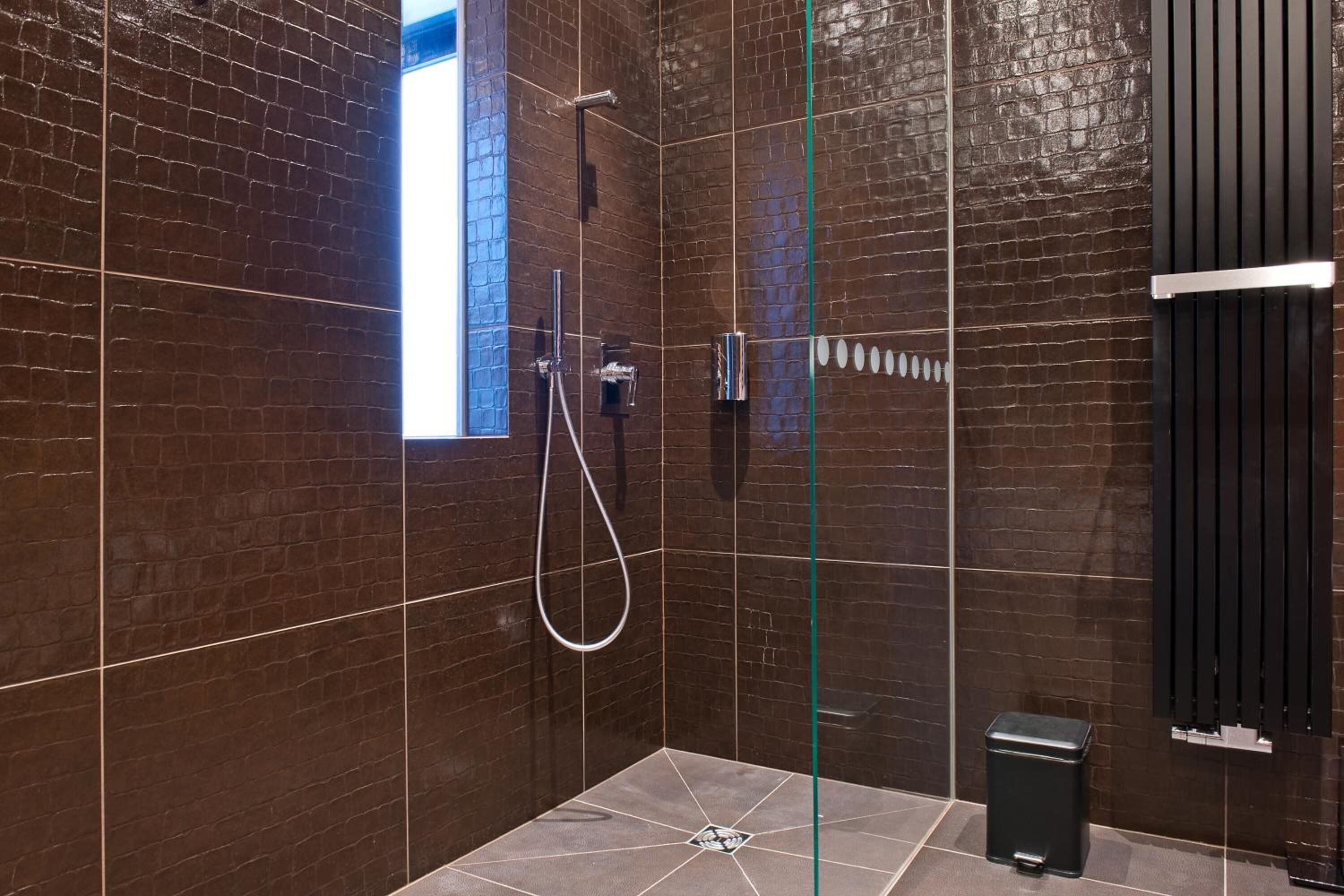 A modern bathroom with dark brown tiled walls, a glass shower enclosure, a handheld showerhead, and a black trash bin.