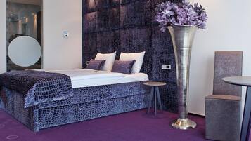 Un dormitorio moderno con un cabecero estampado, un espejo redondo, una cama con ropa de cama blanca, un alto jarrón metálico con flores, una mesita redonda y una silla tapizada de color gris.