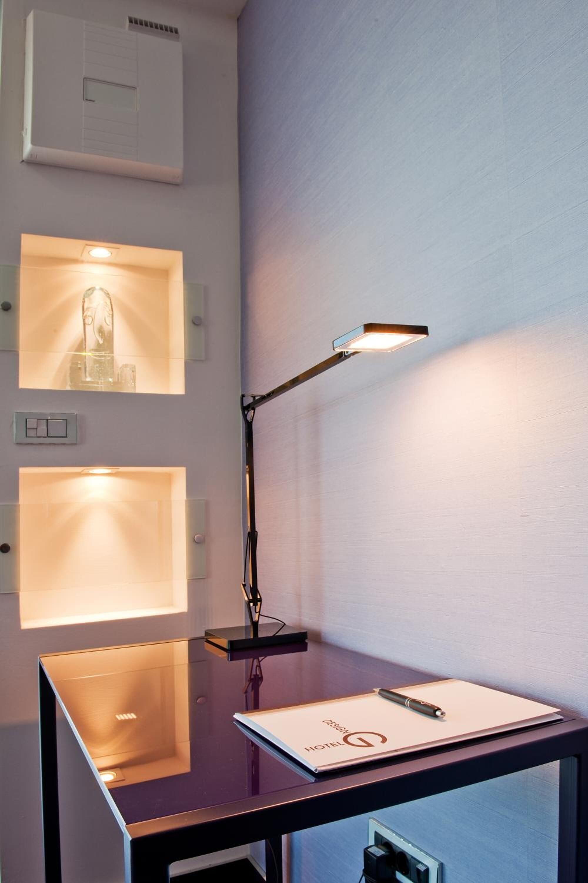 Una scrivania con lampada, un quaderno e una penna. Sono presenti interruttori della luce e un termostato montati a parete sopra la scrivania.