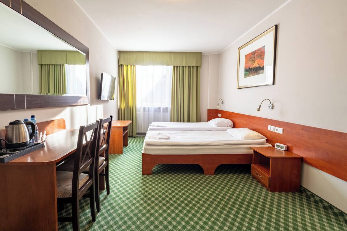Hotellihuone, jossa on sänky, kirjoituspöytä, jossa on vedenkeitin, tuoli, yöpöytä, jossa on kello, ja ikkuna, jossa on verhot.