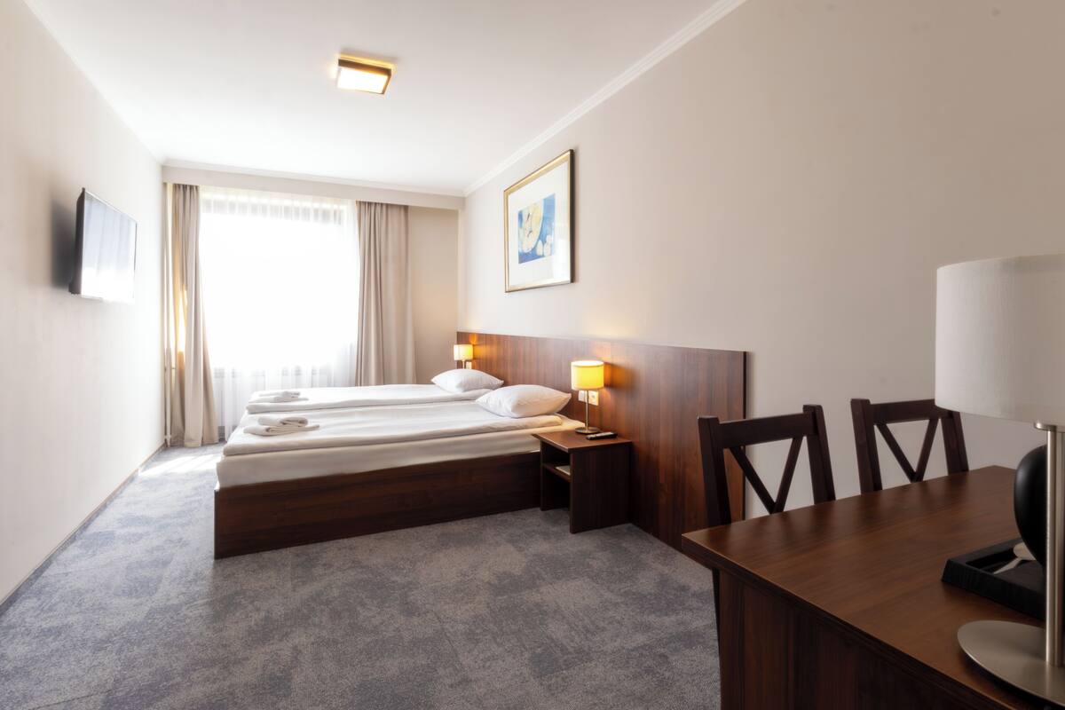 Hotellihuone, jossa on sänky, yöpöytä, lamppu, televisio sekä ruokapöytä tuoleineen.
