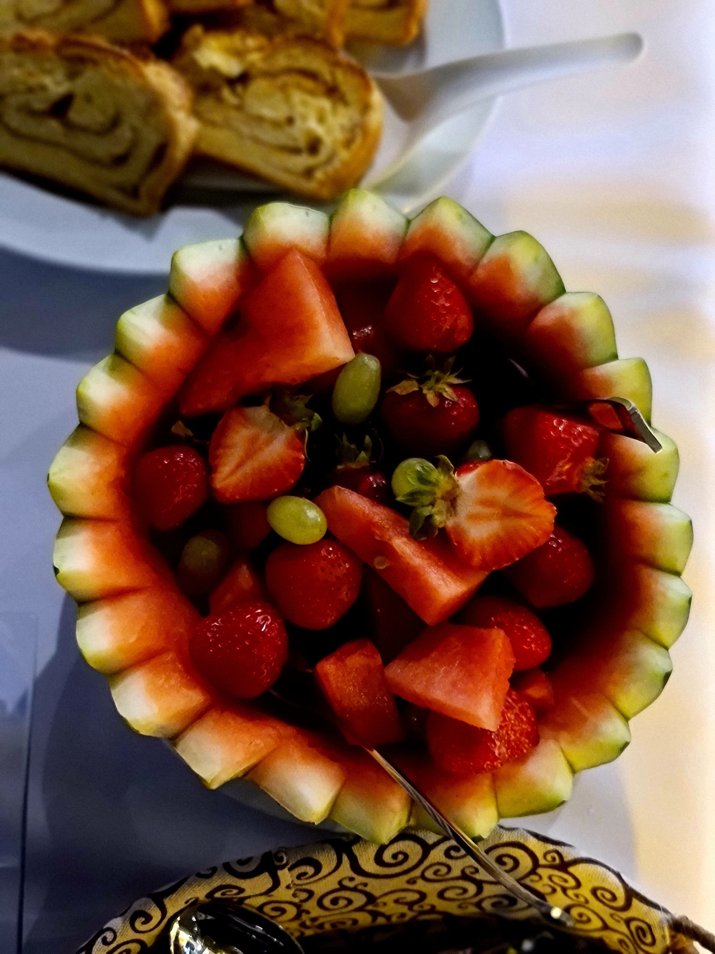 A fruit salad with watermelon, strawberries, and grapes, and a rolled pastry in the background.