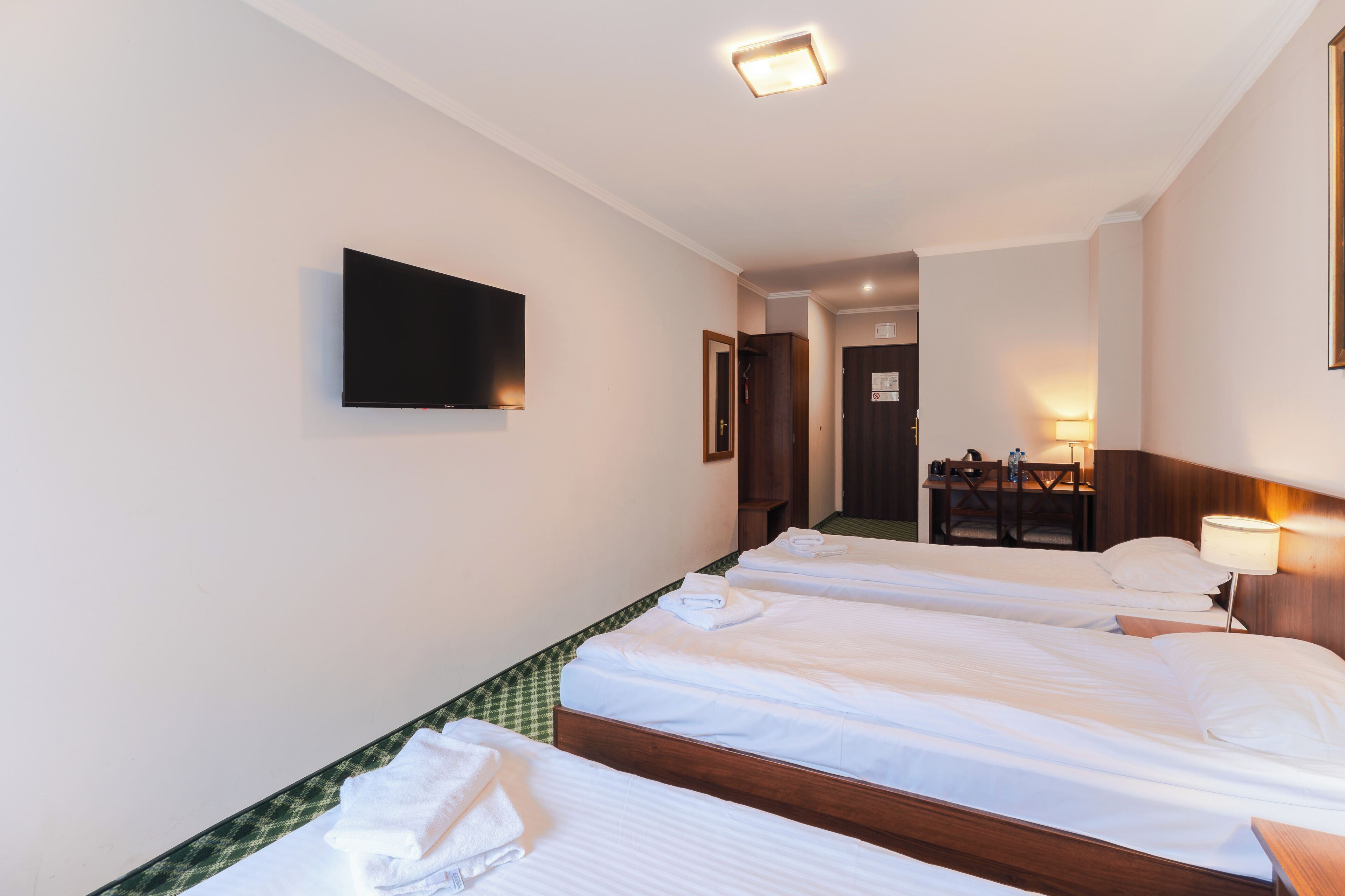 A hotel room with two single beds, a flat-screen TV mounted on the wall, a desk with a chair, and a table lamp.