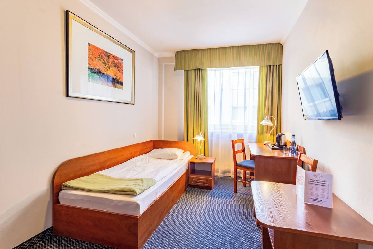 Hotellihuone, jossa on yksi sänky, työpöytä tuolilla, televisio ja ikkuna, jossa on verhot.