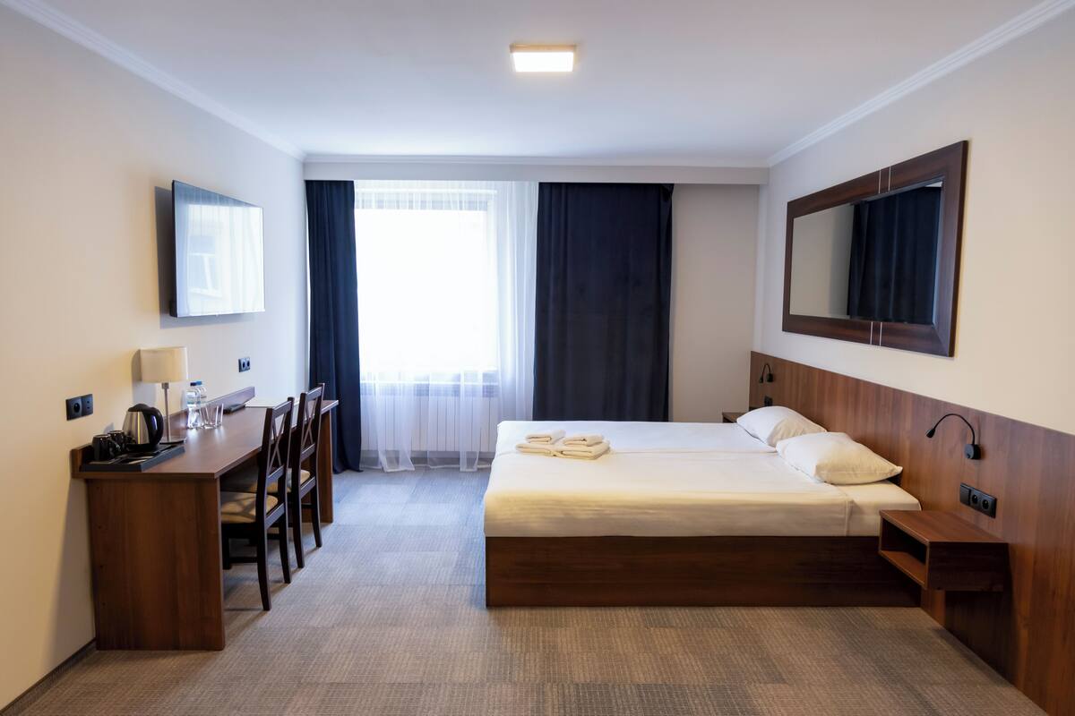 Hotellihuone, jossa on sänky, työpöytä lampun kanssa, tuoli, pieni pöytä, peili ja ikkuna, jossa on verhot.