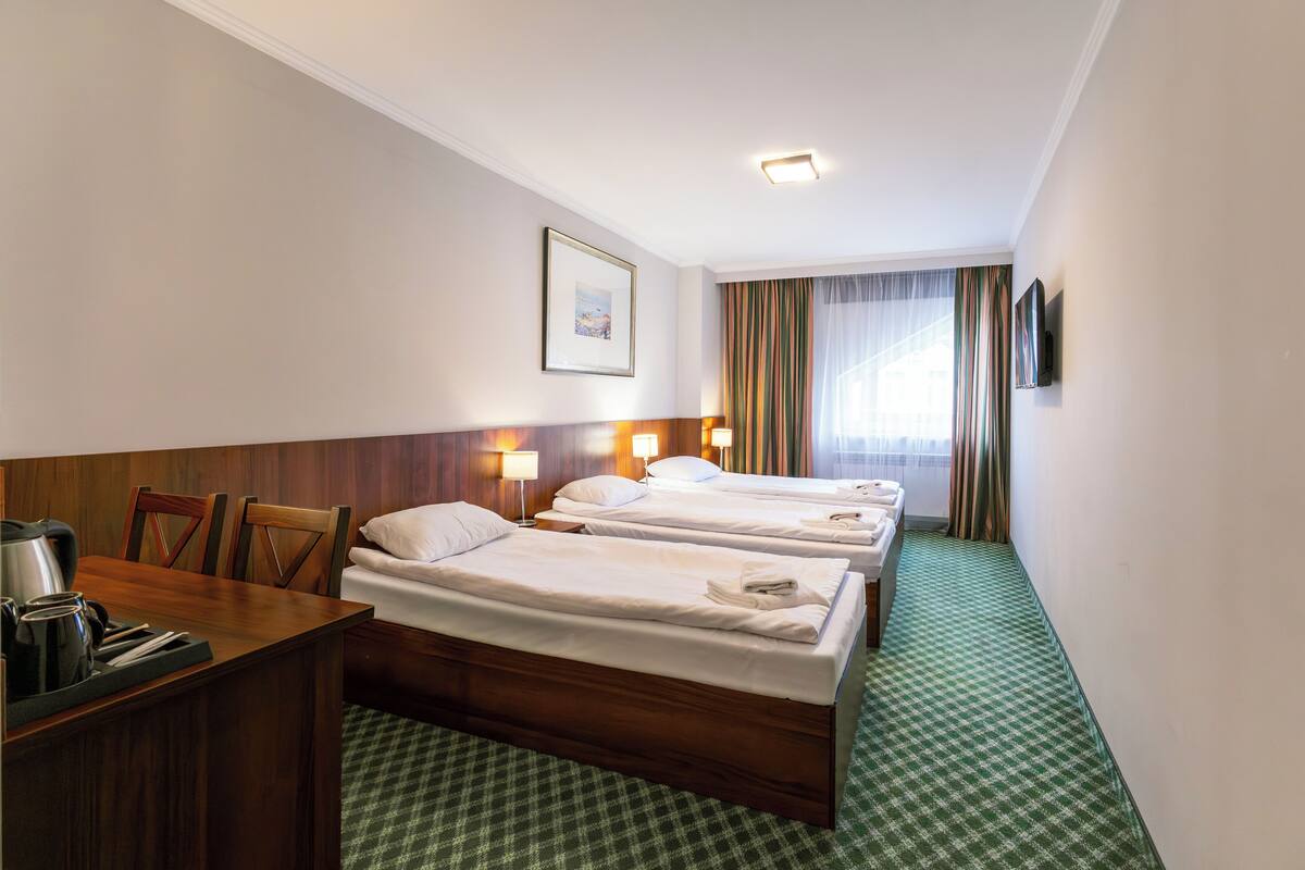 Hotellihuone, jossa on kolme erillistä sänkyä, puinen työpöytä, jossa on vedenkeitin ja kuppeja, seinällä kehystetty taulu ja ikkuna, jossa on verhot.