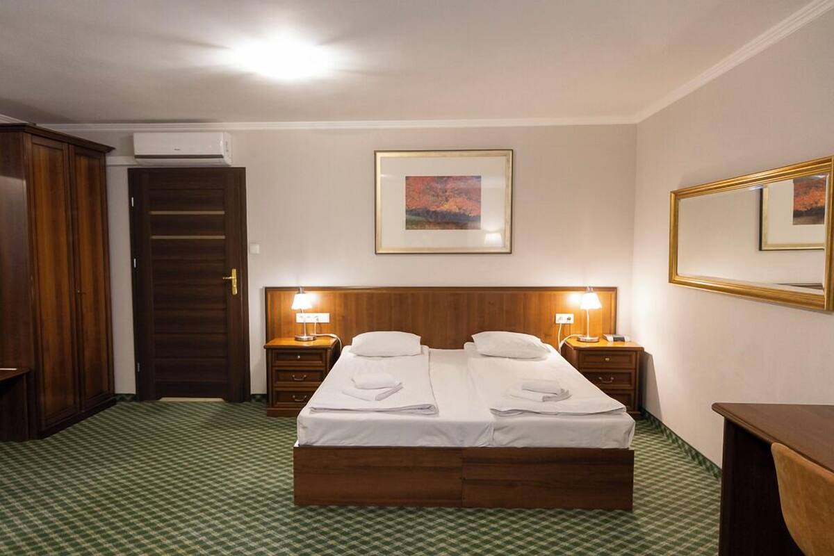 Hotellihuone, jossa on parivuode, kaksi yöpöytää valaisimineen, puinen vaatekaappi, ovi ja seinällä oleva kehystetty taulu.