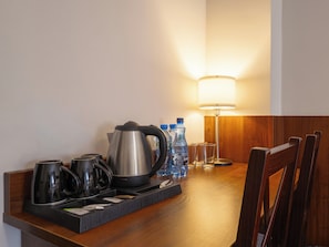 Una habitación de hotel con una mesa de madera, una tetera, tazas, botellas de agua y una lámpara.