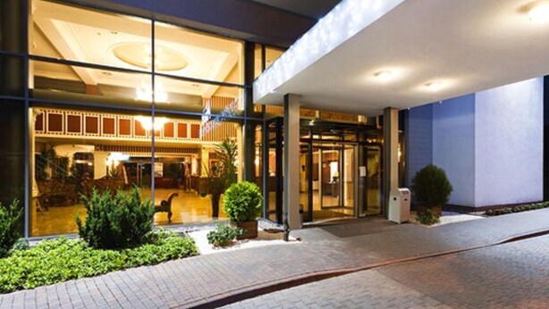 Una entrada de hotel moderna con fachada de vidrio, un espacio exterior cubierto y un camino de ladrillo que conduce a la entrada.