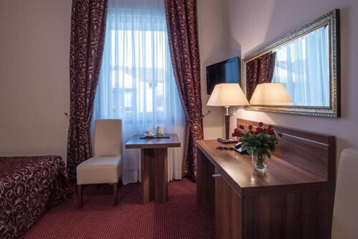 Une chambre d’hôtel avec un lit, une chaise, une petite table avec une lampe, un miroir et un vase avec des fleurs.